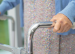 Elderly person gripping the chrome handles of a rollator walker at the front.