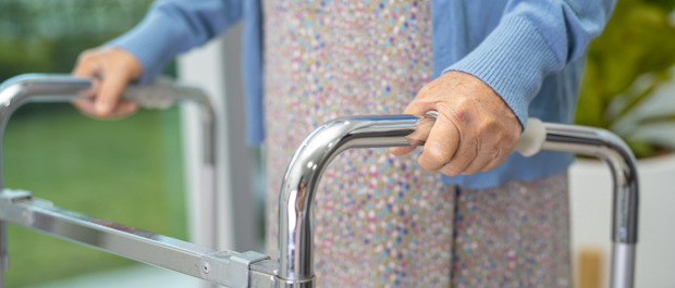 Elderly person gripping the chrome handles of a rollator walker at the front.